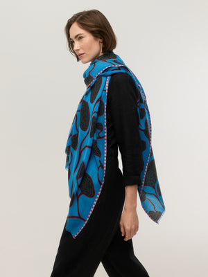 Side view of a bright blue fine merino wool rectangular scarf with black organic botanical print and red stems, pink striped border and frayed edges, draped over the shoulder, Princesse & Dragon