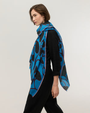 Side view of a bright blue fine merino wool rectangular scarf with black organic botanical print and red stems, pink striped border and frayed edges, draped over the shoulder, Princesse & Dragon