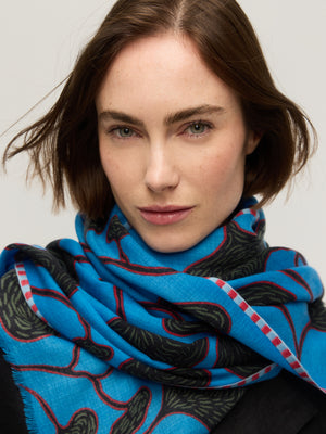 Gros plan sur un foulard en fine laine mérinos bleu vif à motif végétal aux formes organiques noires et tiges rouges, bordure rayée rose, enroulé autour du cou, Princesse & Dragon