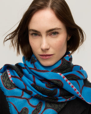 Gros plan sur un foulard en fine laine mérinos bleu vif à motif végétal aux formes organiques noires et tiges rouges, bordure rayée rose, enroulé autour du cou, Princesse & Dragon