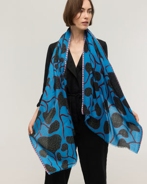 Foulard rectangulaire en fine laine mérinos bleu vif à motif végétal aux formes organiques noires et tiges rouges, bordure rayée rose et bords frangés, porté ouvert sur les épaules, Princesse & Dragon